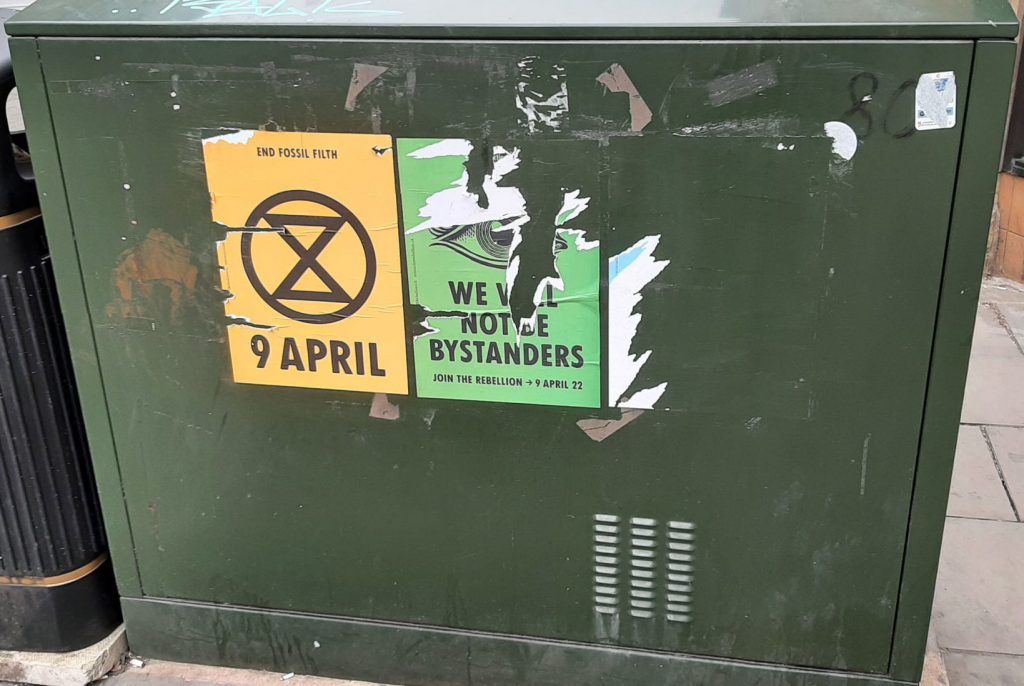 A green metal box (housing cables) with three protest posters pasted on it. Of which all three have been ripped, with one almost ripped off. 